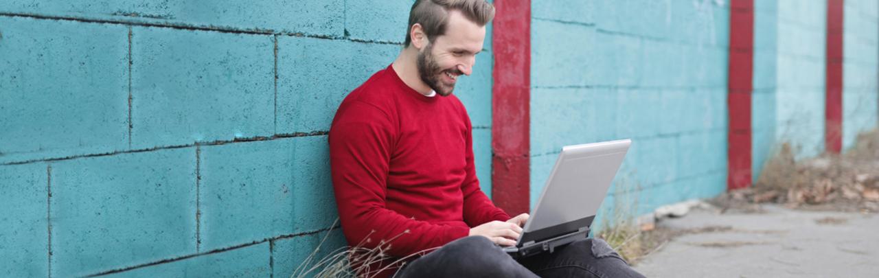 Male smiling while working on laptop sitting on ground outside 