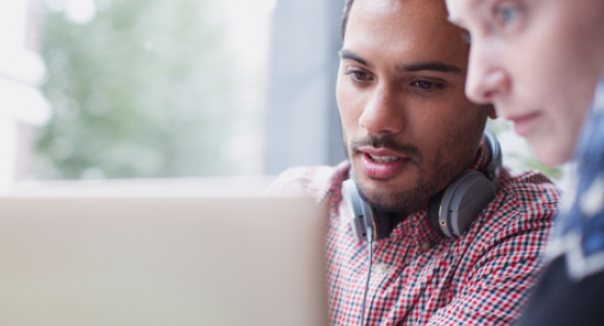 Man wearing headphones viewing a laptop screen while seated next to another person.