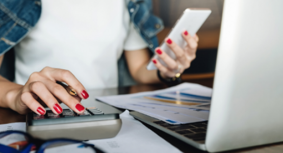 Female seated using a laptop computer and a 10-key while viewing her phone.